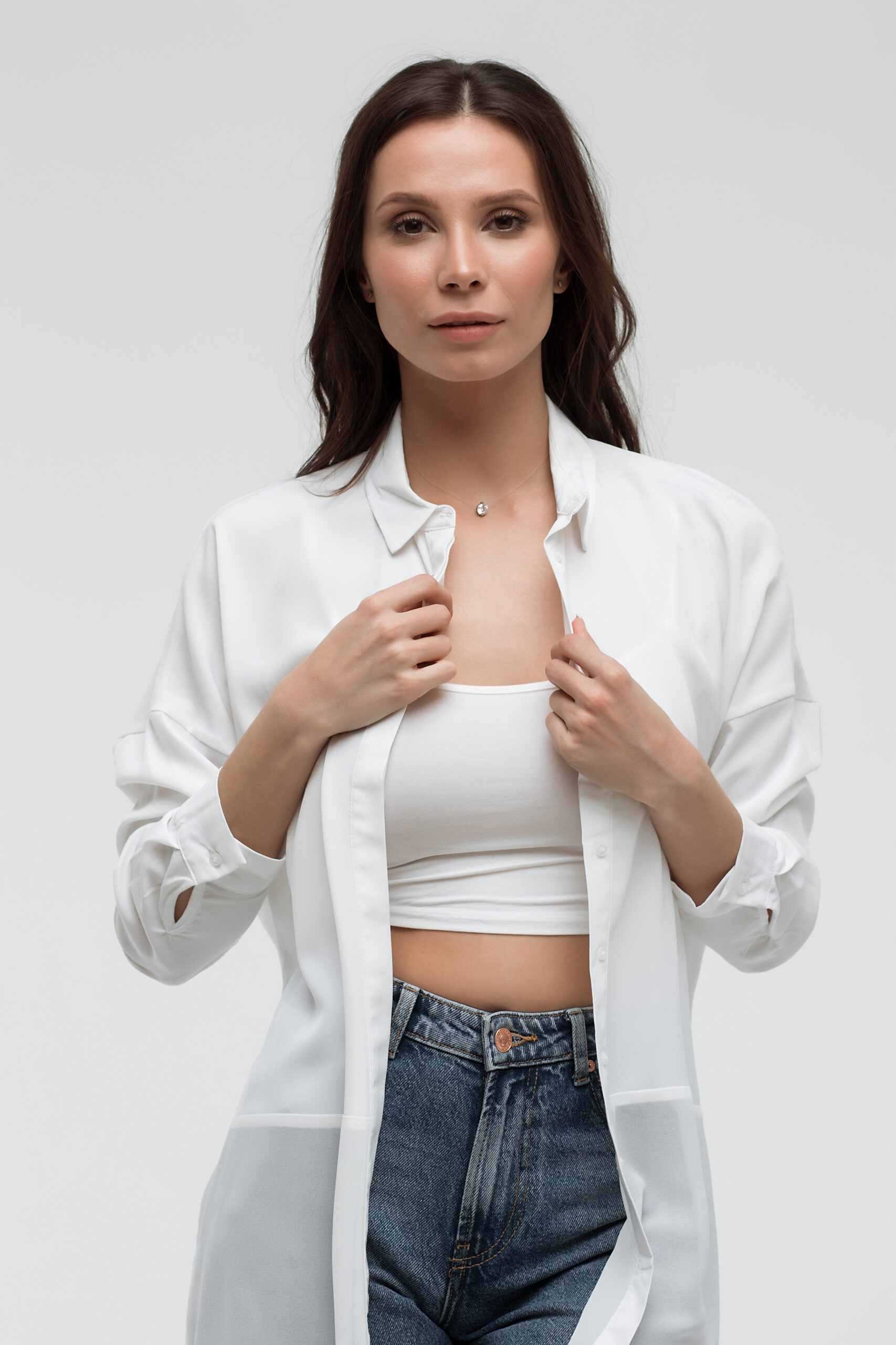 Woman in white shirt and jeans looks confidently at the camera. She holds the shirt slightly open, revealing a white cropped tank top. Neutral background.